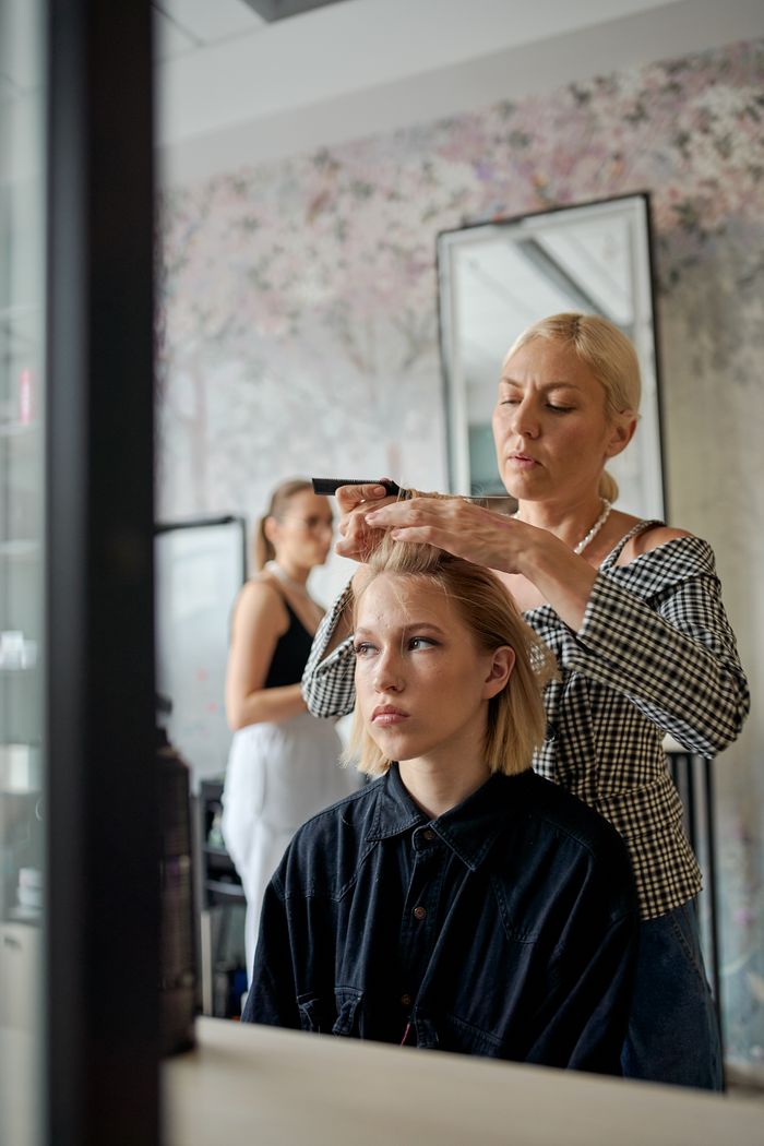 Adult female hairstylist straightening hair of dreamy client with iron reflecting in mirror in beauty salon